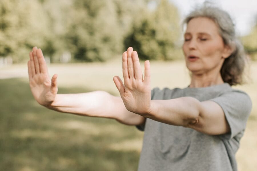 Oudere vrouw doet een rustige qi gong-oefening met gespreide armen en gesloten ogen in een zonnig park