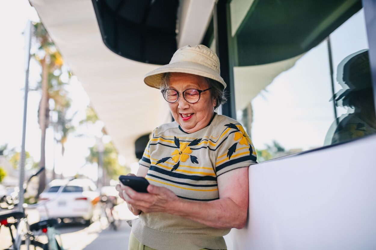 Lachende oudere vrouw die buiten in de zon een bericht typt op haar smartphone