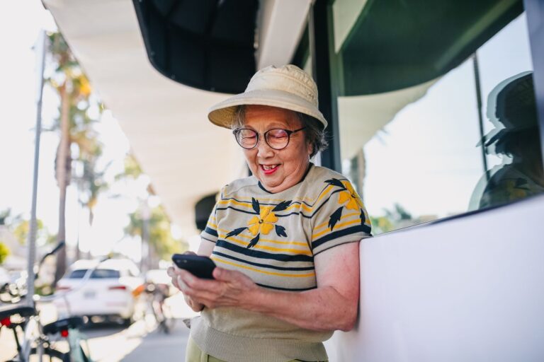 Lachende oudere vrouw die buiten in de zon een bericht typt op haar smartphone