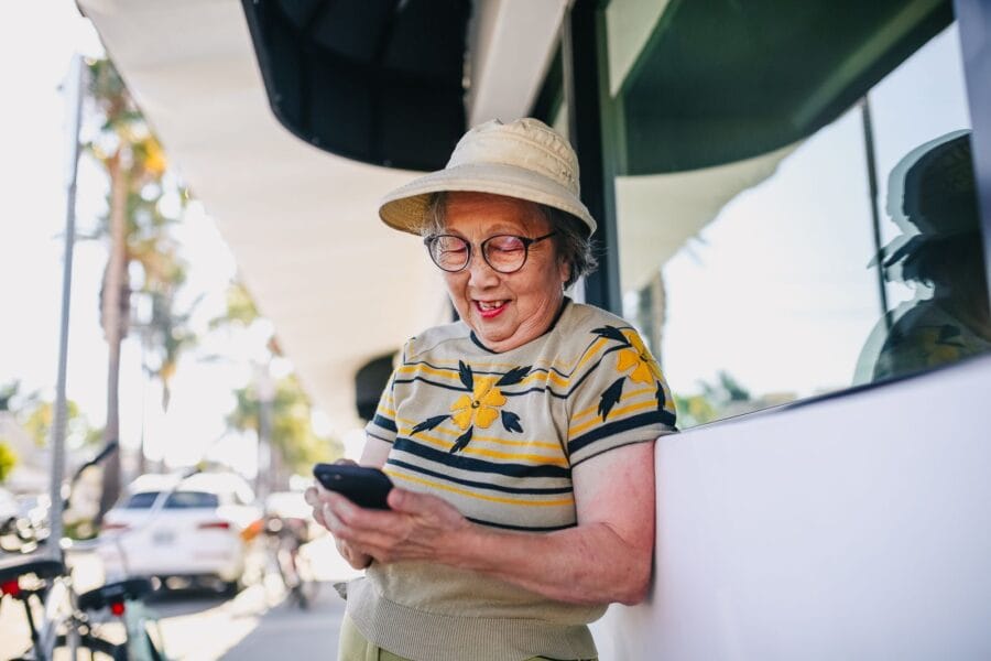 Lachende oudere vrouw die buiten in de zon een bericht typt op haar smartphone